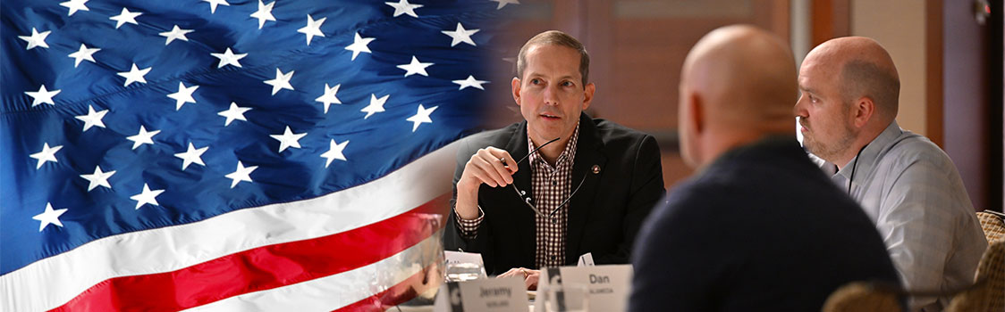 Group seated around a conference table with nameplates visible, engaged in discussion. A large American flag is prominently displayed on the left side of the image.