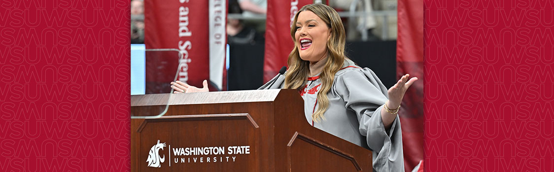 Alumna Jamie Kern Lima speaking at a podium during the Carson College spring commencement ceremony. The podium displays the Washington State University logo, and red banners with WSU branding are visible in the background.