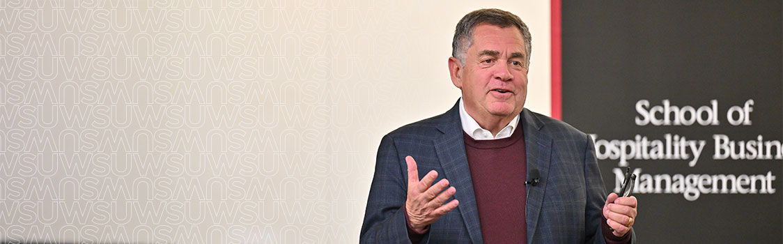 John Oppenheimer giving a presentation in front of a wall with repeated WSU branding and a black panel displaying the text “School of Hospitality Business Management.” The individual is wearing a dark plaid jacket over a maroon sweater and holding a presentation remote.