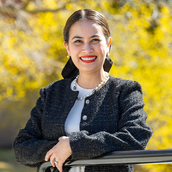 Mia Darnell wearing a dark textured jacket and light top, standing outdoors with bright yellow foliage in the background.