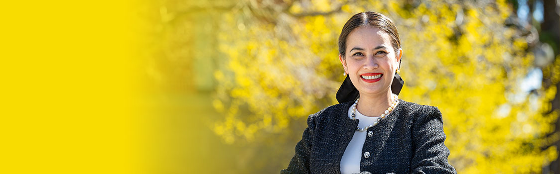 Mia Darnell wearing a dark textured jacket and light top, standing outdoors with bright yellow foliage in the background and a solid yellow gradient on the left side of the image.