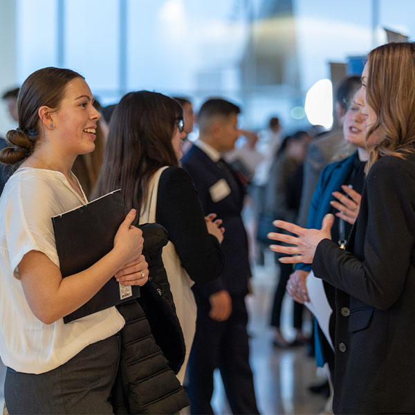 People engaged in conversation at a networking event, holding folders and wearing name tags, with others mingling in the background.