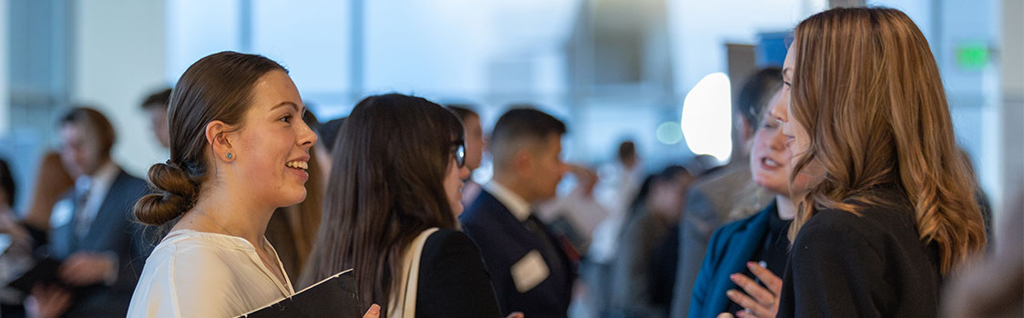 People engaged in conversation at a networking event, holding folders and wearing name tags, with others mingling in the background.
