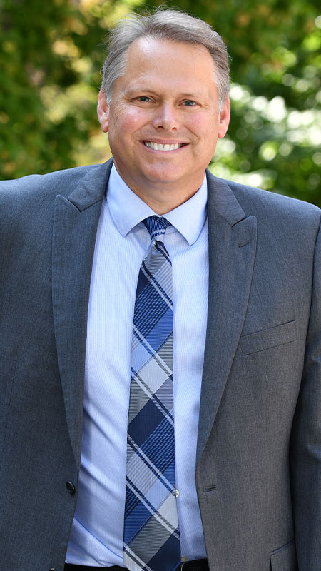 PhD Director Chuck Munson wearing a gray suit, light blue shirt, and patterned blue tie, standing outdoors with green foliage in the background.