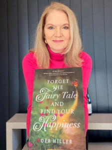 Deb Miller holds her book titled “Forget the Fairy Tale and Find Your Happiness” in front of a dark paneled background.