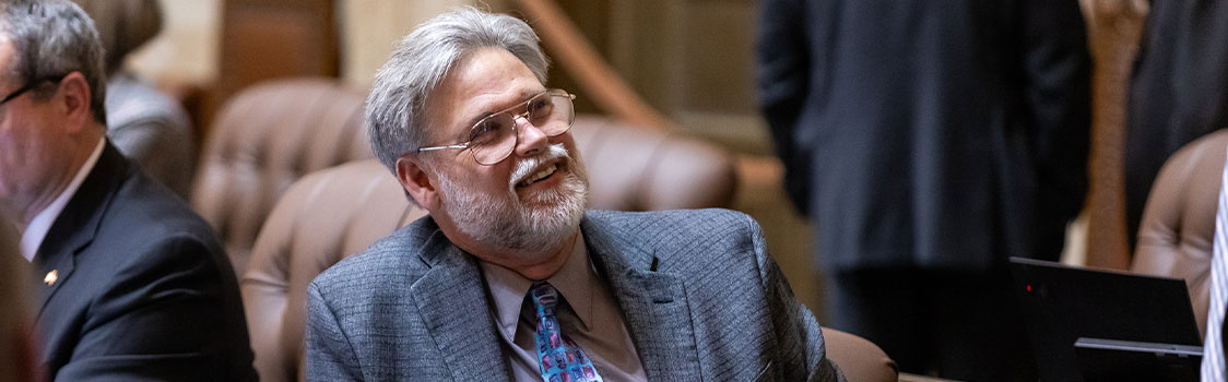 Representative Ed Orcutt seated in a legislative chamber, wearing a gray patterned suit and colorful tie. Other attendees and leather chairs are visible in the background, along with a laptop on a desk.