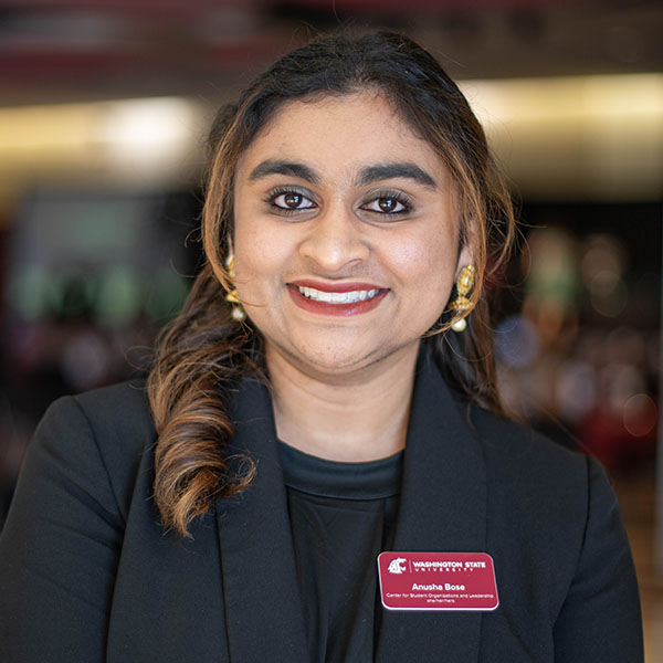Anusha Bose wearing a black blazer and gold earrings, with a name badge that reads “Anusha Bose” at a professional event.