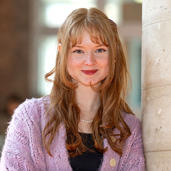 Abby Vance wearing a lavender knit sweater and a pearl necklace, standing indoors with a softly blurred background.