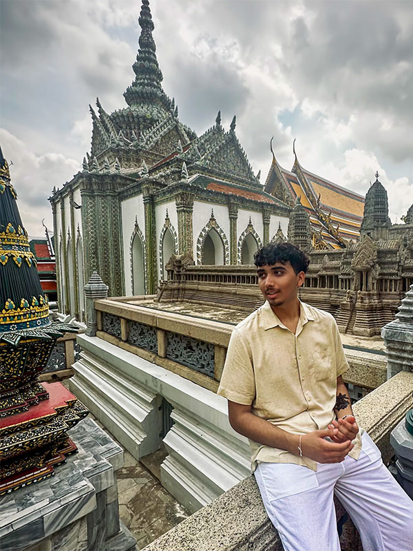 Jose Lopez sits on a stone railing at the Grand Palace in Bangkok, Thailand, with ornate temple spires and intricate architecture in the background under a cloudy sky.