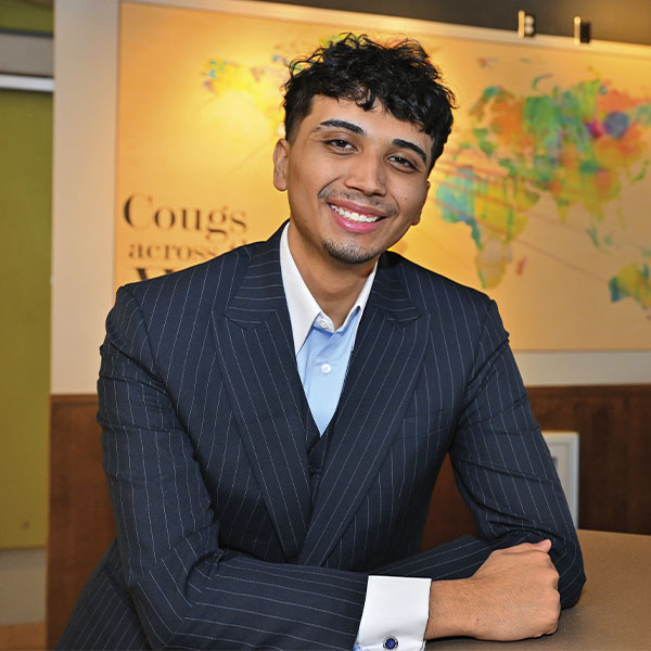 Jose Lopez wearing a dark pinstripe suit and light blue shirt, seated at a table with arms resting on the surface. A colorful world map and the words “Cougs across the world” are visible on the wall in the background.