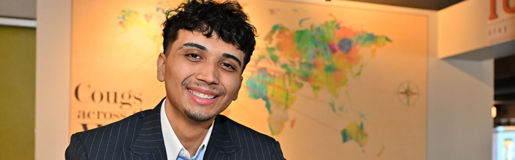Jose Lopez wearing a dark pinstripe suit and light blue shirt, seated at a table with arms resting on the surface. A colorful world map and the words “Cougs across the world” are visible on the wall in the background.