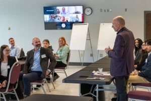 A speaker addresses participants seated around tables during the Tri-Cities One Hanford Leadership Academy, with flip charts and a video conference displayed on a wall monitor.
