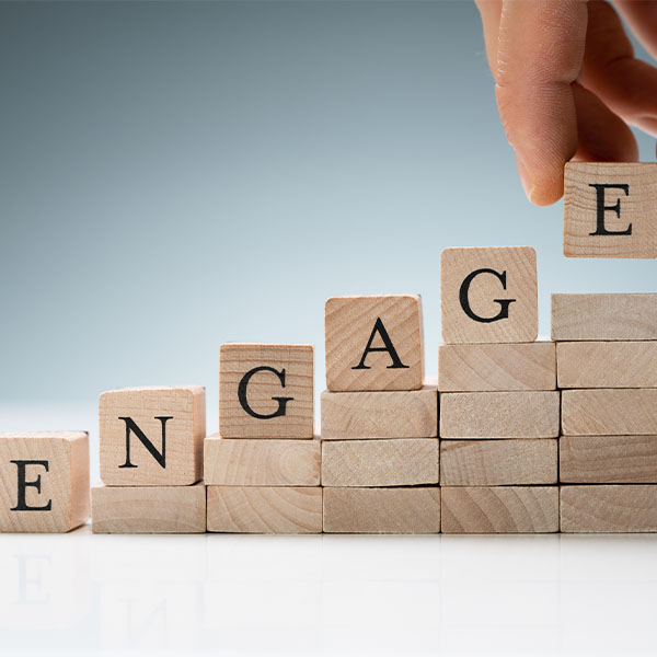 Stacked wooden blocks spelling the word “ENGAGE,” arranged in ascending steps, with a hand placing the final block with the letter “E” on top.