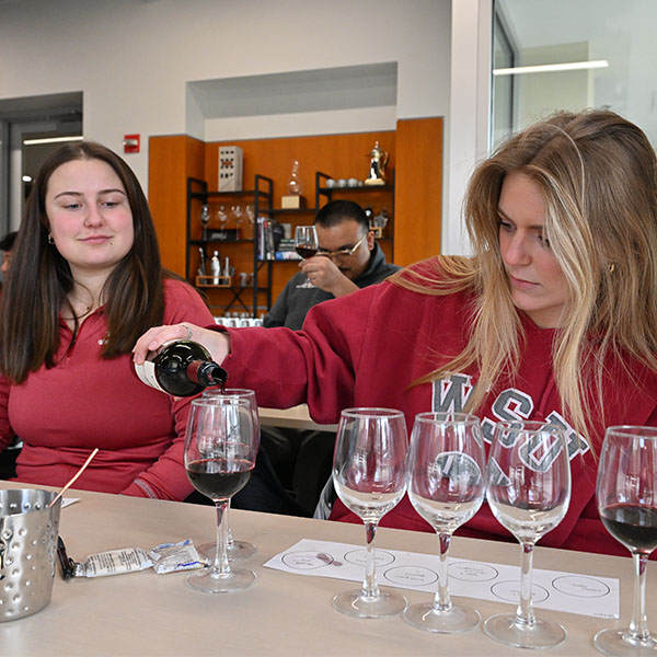 Students wearing a WSU sweatshirts pour red wine into tasting glasses during a wine tasting session, with multiple glasses and a tasting sheet on the table.