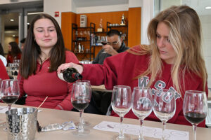 Students wearing a WSU sweatshirts pour red wine into tasting glasses during a wine tasting session, with multiple glasses and a tasting sheet on the table.