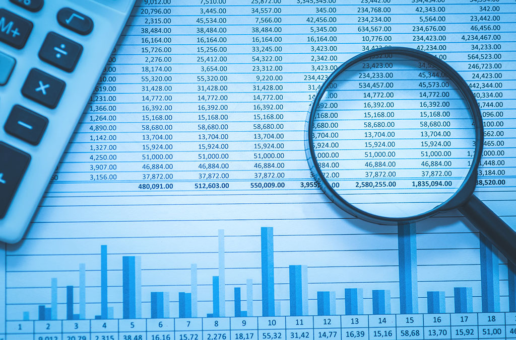 Close-up of a financial spreadsheet with rows of numbers, a magnifying glass highlighting figures, and a calculator partially visible on the side. A bar chart appears at the bottom of the page.