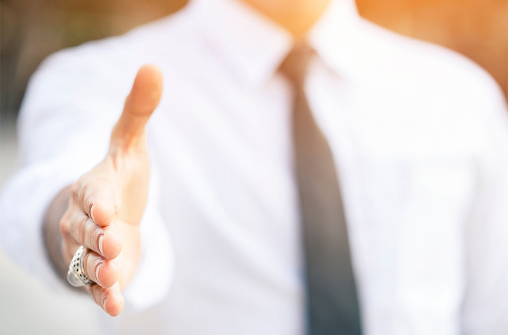 Person in a white dress shirt and black tie extending a hand forward for a handshake.