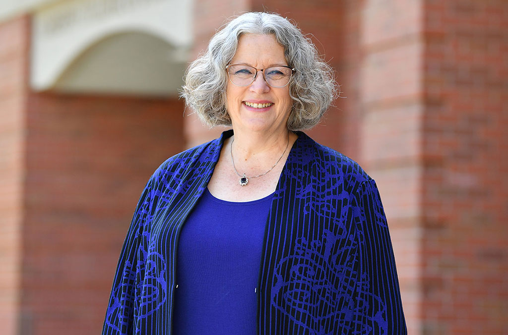 Dean Debbie Compeau standing outdoors in front of a brick building, wearing a blue top with a patterned jacket and a pendant necklace.