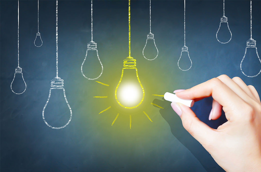 Hand drawing a glowing yellow light bulb with chalk on a dark board, surrounded by outlines of other light bulbs.
