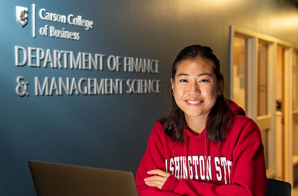 Person wearing a red Washington State University hoodie seated at a desk with a laptop, in front of a wall sign reading “Carson College of Business, Department of Finance & Management Science.”