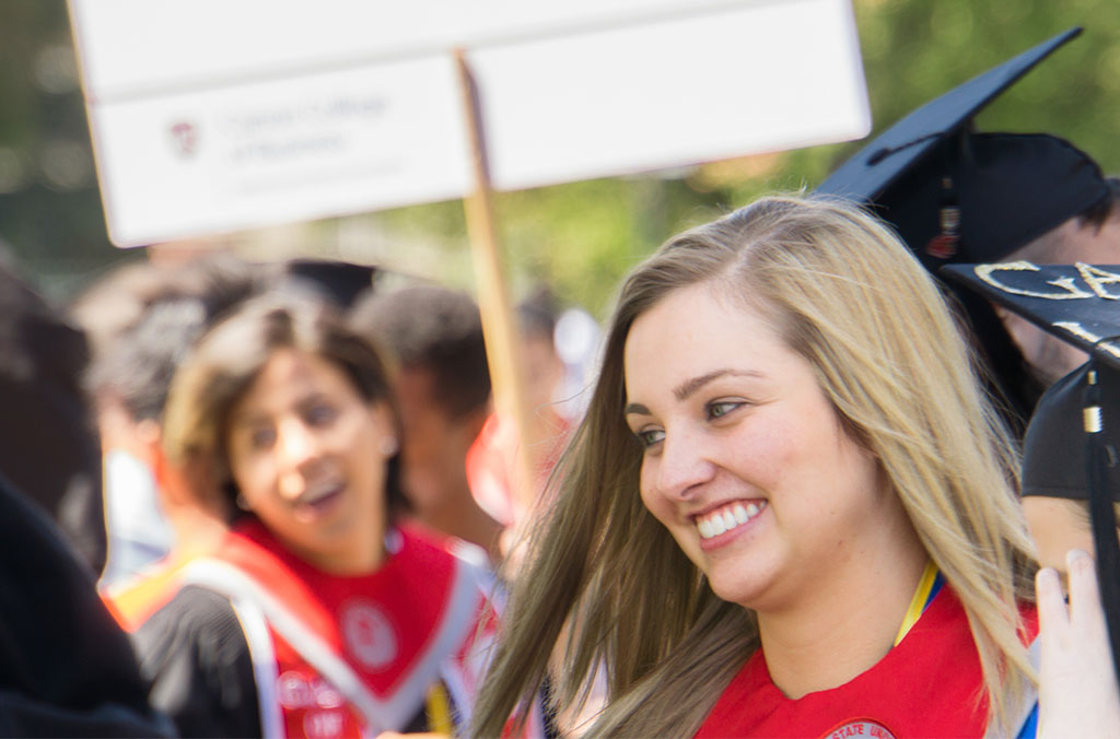 Graduates in caps and gowns walking in a procession, with a sign visible in the background.