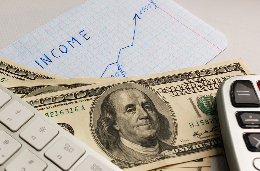 Keyboard, calculator, and U.S. hundred-dollar bills placed on a desk with a graph labeled “Income” showing an upward trend.