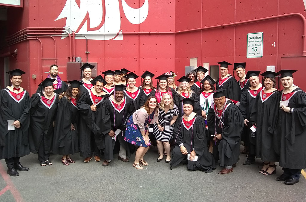 Large group of graduates in black caps and gowns posing together in front of a red wall with the Washington State University logo.