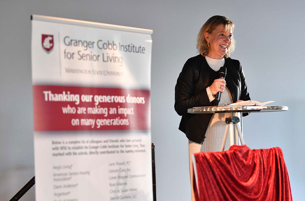 Person standing at a podium holding a microphone, next to a banner that reads “Granger Cobb Institute for Senior Living, Washington State University” and “Thanking our generous donors who are making an impact on many generations.”