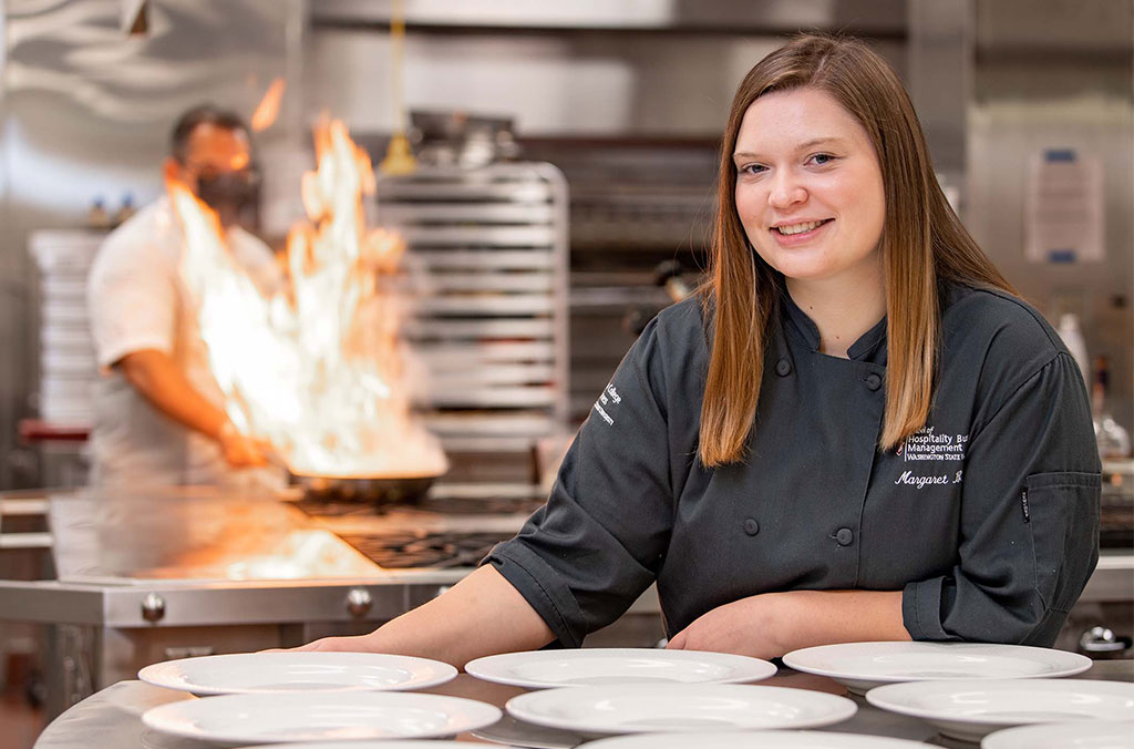 Person in a professional kitchen wearing a dark chef coat, standing behind a row of white plates, while another person cooks at a stove with flames rising from a pan in the background.