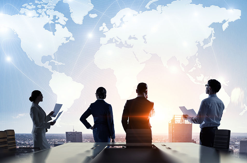 Four business professionals standing in a modern office, reviewing documents while looking out at a city skyline with a large world map and connected network lines displayed in the background.