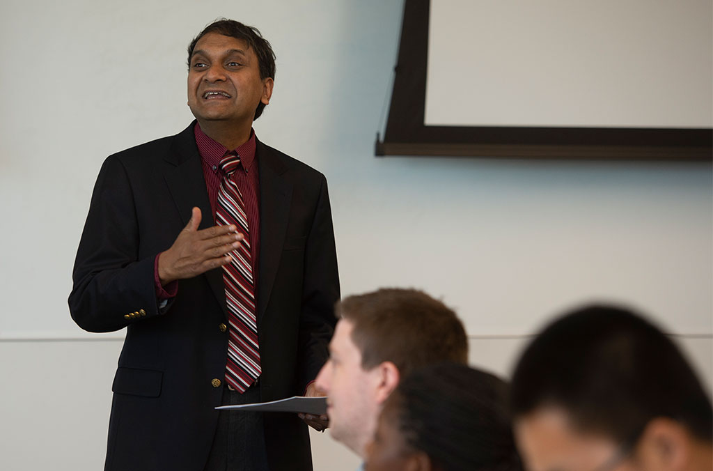Person in a dark suit and striped tie speaking in front of a projection screen, holding papers, with others seated in the foreground.