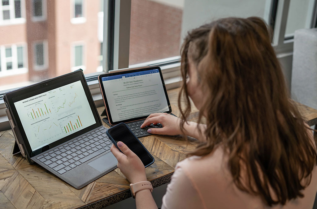 Person seated at a desk using two tablets and a smartphone, with charts and graphs displayed on one screen and text on the other.