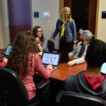 Thumbnail of http://Group%20of%20students%20seated%20around%20a%20conference%20table%20with%20laptops%20open,%20while%20an%20instructor%20stands%20at%20the%20head%20of%20the%20table.%20A%20projection%20screen%20displays%20spreadsheet%20data%20in%20the%20background.