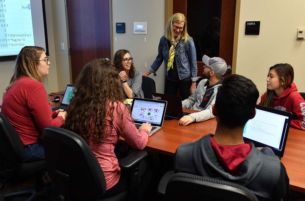 Group of students seated around a conference table with laptops open, while an instructor stands at the head of the table. A projection screen displays spreadsheet data in the background.