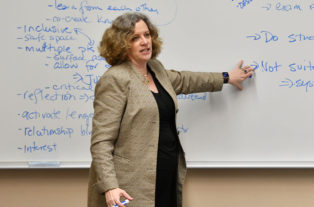 Debbie Compeau standing in front of a whiteboard covered with handwritten notes, pointing to text while holding a marker.