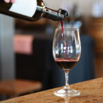 Thumbnail of http://Red%20wine%20being%20poured%20from%20a%20bottle%20into%20a%20clear%20wine%20glass%20on%20a%20polished%20countertop,%20with%20wooden%20barrels%20blurred%20in%20the%20background.