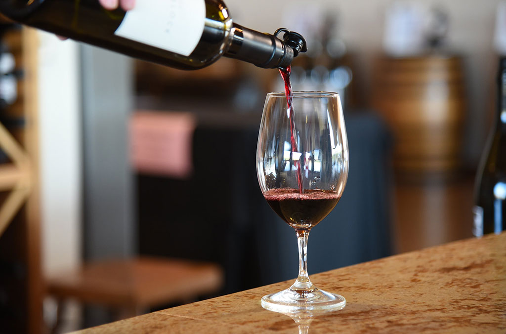 Red wine being poured from a bottle into a clear wine glass on a polished countertop, with wooden barrels blurred in the background.