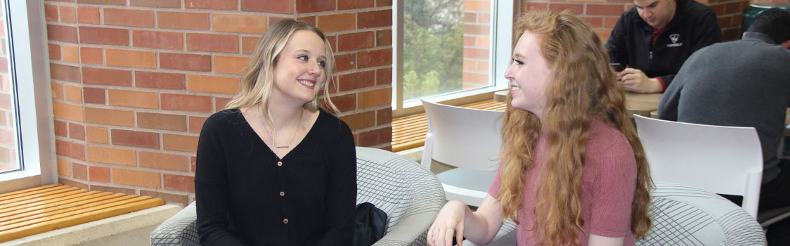 Two students sitting in lounge chairs and talking in a campus common area with large windows and brick walls.