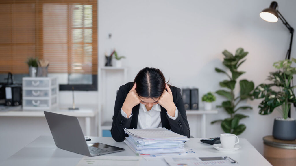 Employee sitting at a desk with head down over a stack of papers, appearing stressed in a workplace setting.