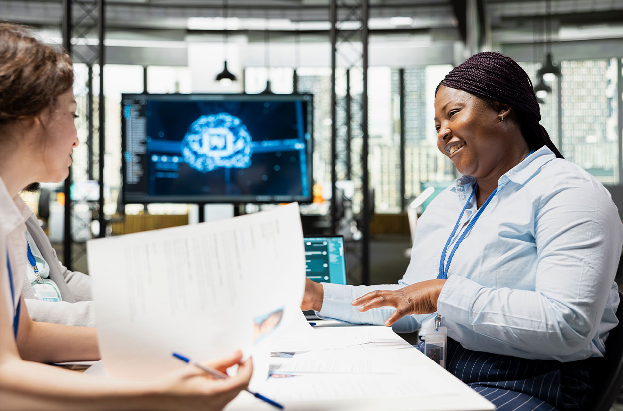 Two professionals sit at a table reviewing printed documents during a discussion, with laptops open and a large screen behind them displaying a digital brain graphic in a modern office setting.