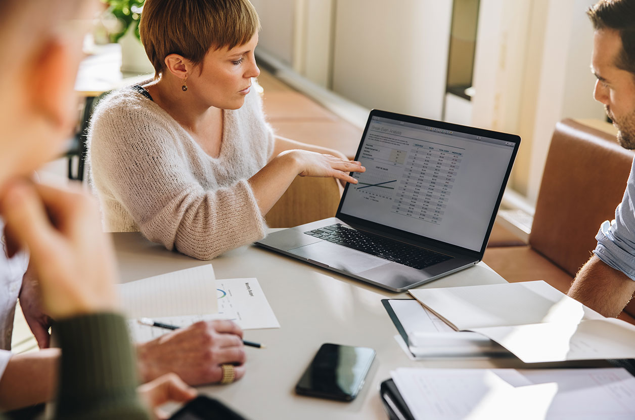 Small group gathers around a table as one person points to a spreadsheet and chart displayed on a laptop during discussion.