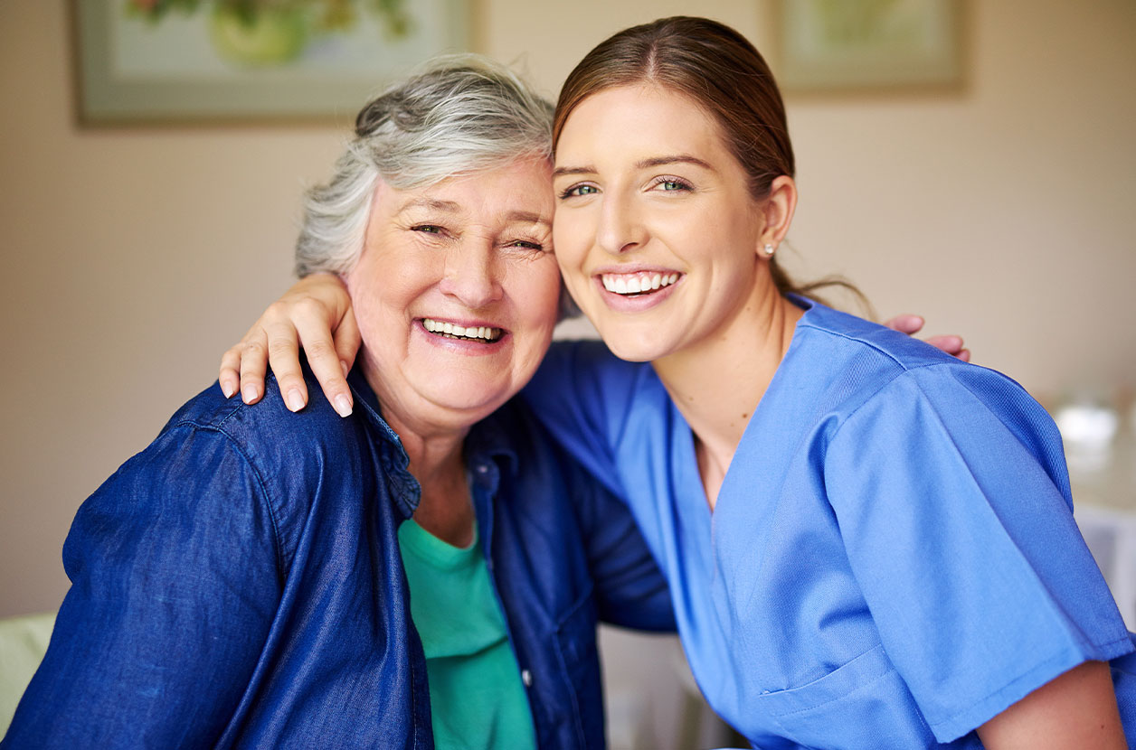 Caregiver in blue scrubs sits beside an older adult, with an arm around their shoulders in a supportive pose.