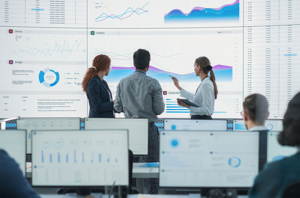 Team reviews wall‑sized dashboards of charts and graphs, pointing to trends in a modern analytics workspace.