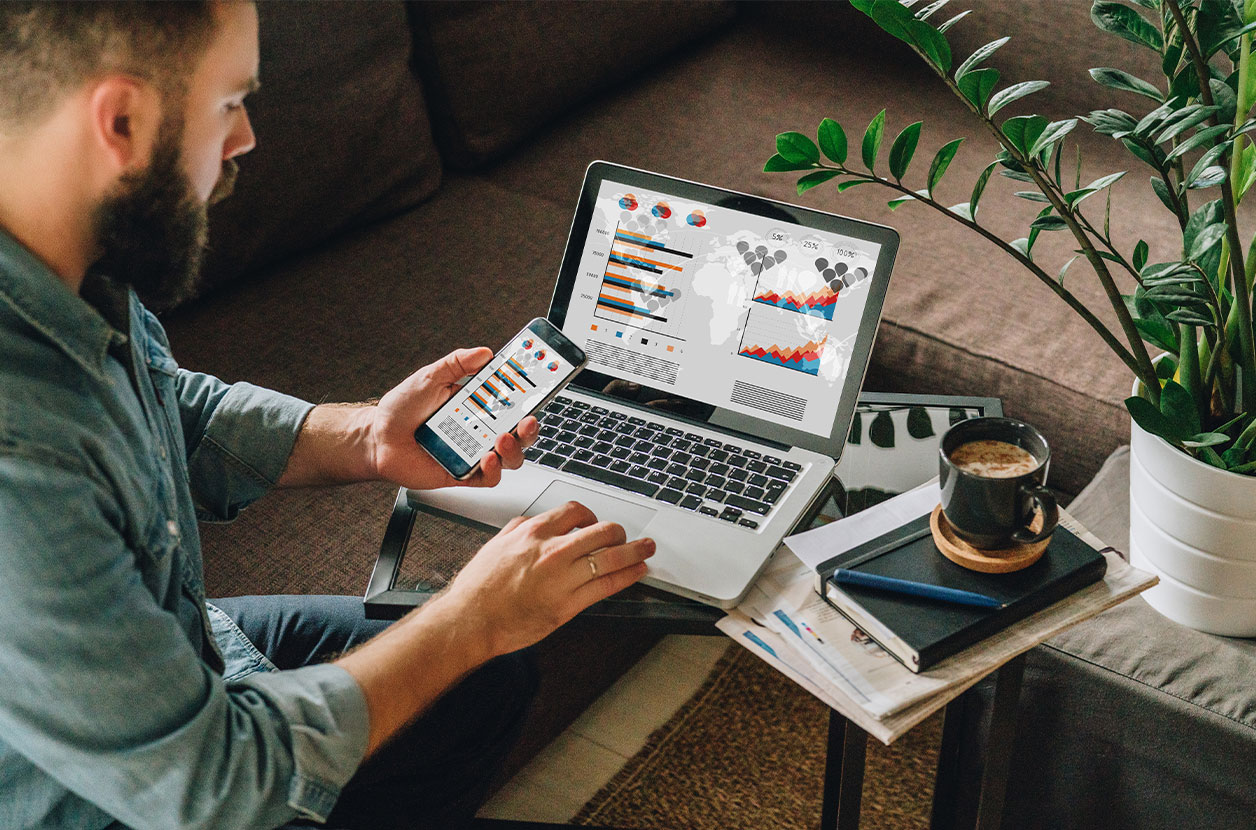 Person checks analytics charts on a laptop and smartphone while seated on a couch, with a notebook, coffee mug, and plant nearby.