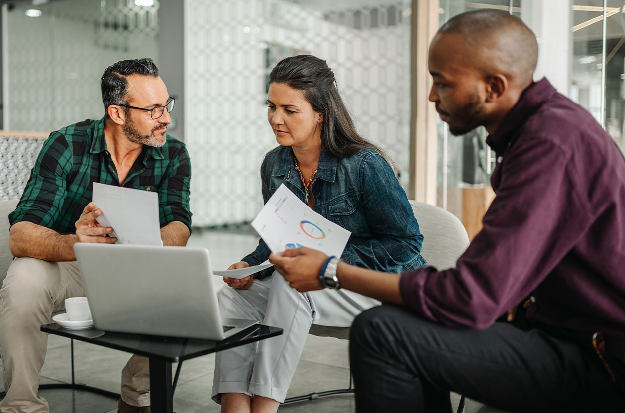Three people sit in a meeting area reviewing printed charts and a laptop, discussing financial data around a small table.