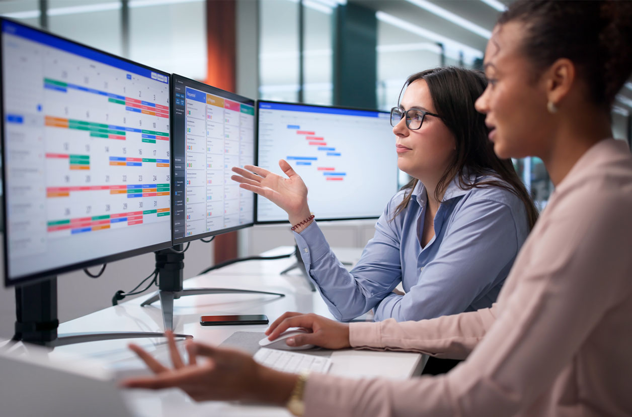Two people discuss project schedules while reviewing task timelines on multiple monitors at a shared workstation.