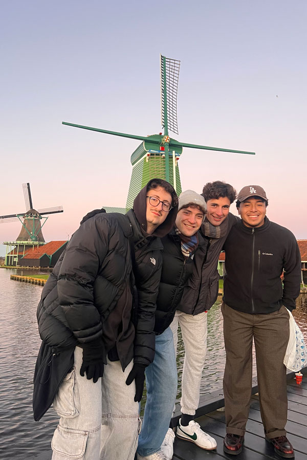 Group of friends pose in front of traditional Dutch windmills near water during a day trip to Amsterdam, with Roberto Muñoz positioned at the far right.