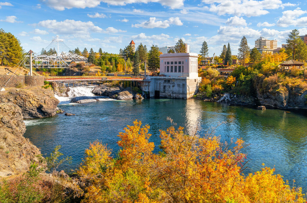 Spokane River flows past a pedestrian suspension bridge and historic building in Riverfront Park, with autumn trees and skyline in Spokane, Washington.