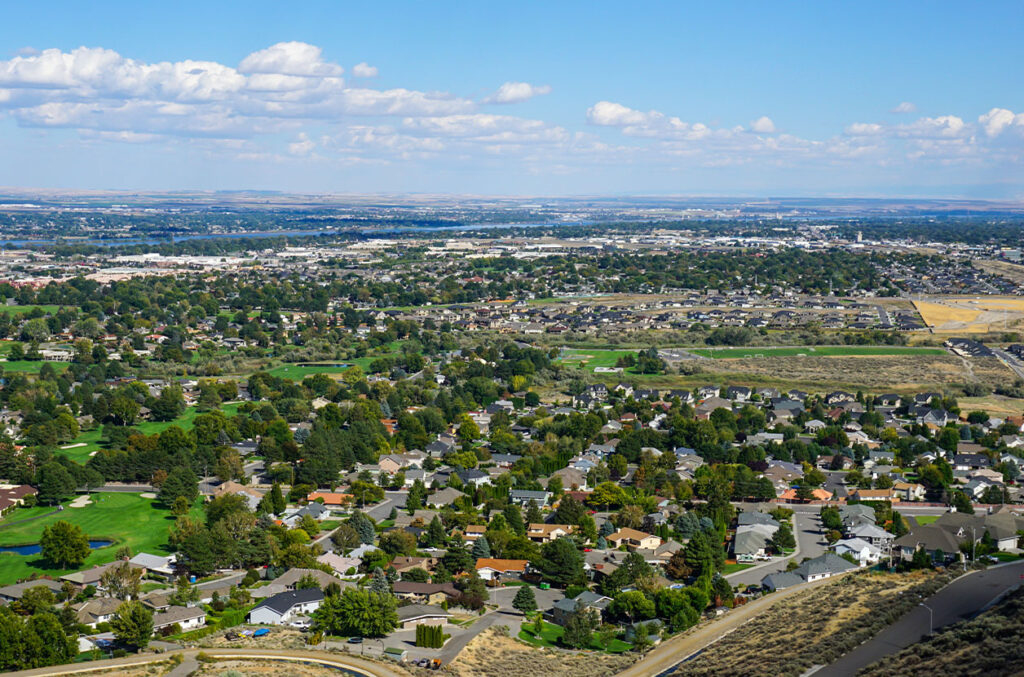 Aerial view of the Tri‑Cities, Washington, showing suburban neighborhoods, green spaces, and the Columbia River stretching across the landscape under a blue sky.