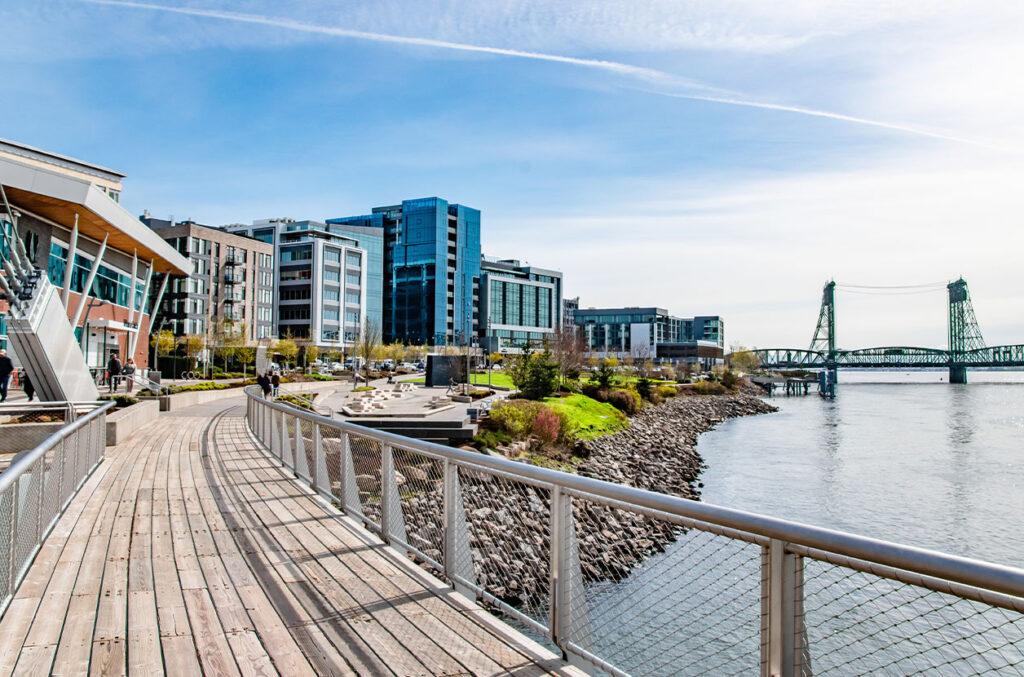 Riverfront walkway in Vancouver, Washington, curves past modern buildings along the Columbia River, with the Interstate Bridge in the distance.